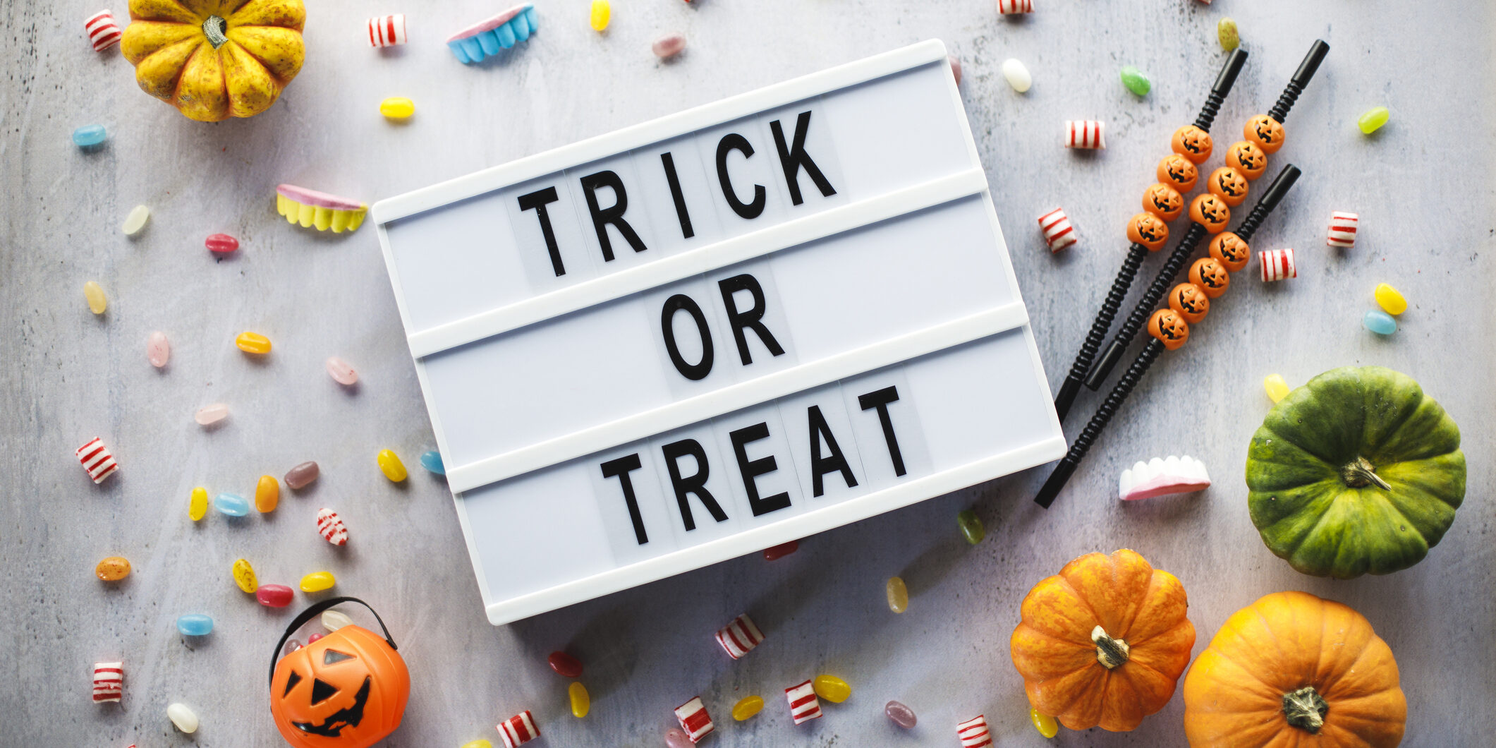 Flat lay of a letter board that says ''trick or treat'', little Jack o' lantern bucket, scattered candy, fun drinking straws and colorful gourds arranged on a white background.