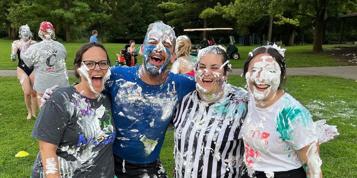 Four young adults laugh with whipped cream on their faces and clothes at MDA Summer Camp