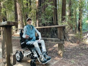 Leslie Krongold drives a power wheelchair on a boardwalk path through a redwood forest at Hendy Woods State Park.
