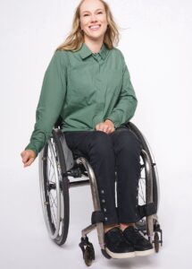 A woman, pictured waist down sitting in a wheelchair wearing pants with snaps down the side of the leg. She is wearing black slip-on shoes. 
