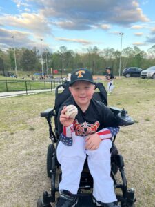 Eight-year-old Bentlee sits in a power wheelchair wearing a baseball uniform and holding up a baseball. He is surrounded by the green grass and black fencing of a ballpark, with a blue sky and white clouds overhead.