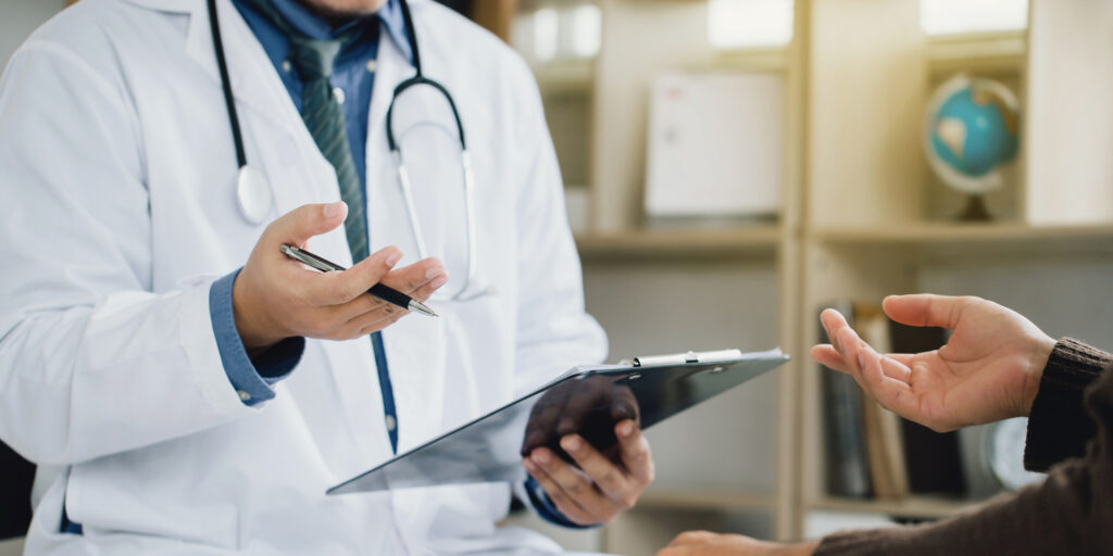 A doctor holds a clipboard and pen while talking to a patient out of the shot