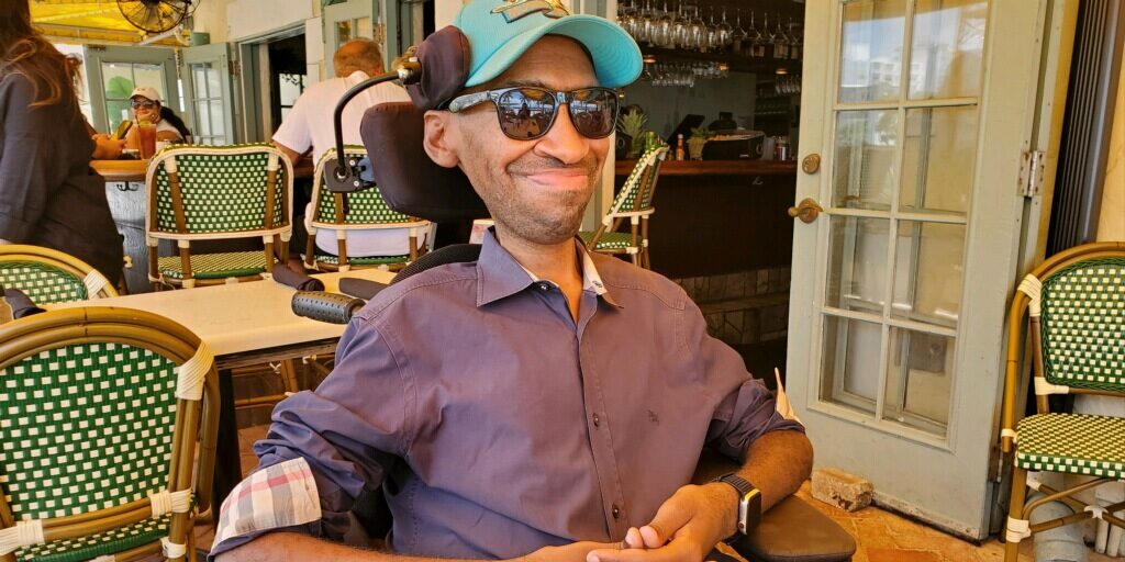 A Black male wearing a teal baseball hat, purple button down shirt, and sunglasses, smiles in his power wheelchair at an outdoor restaurant