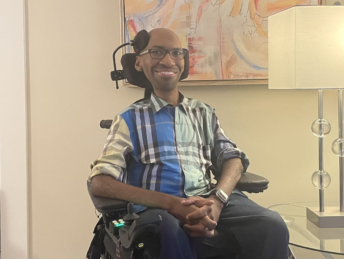 A Black man in a plaide shirt sits in a power wheelchair next to a lamp