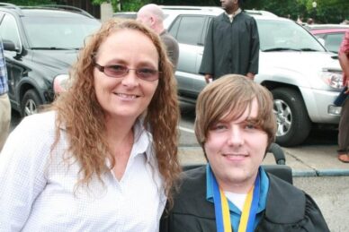 A young man in a graduation gown and power wheelchair smiles with his mother