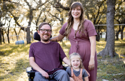 A man in wheelchair wearing glasses and a purple shirt smiles next to his wife in a purple dress and young daughter