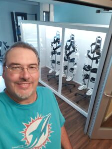 Selfie of Dwayne Wilson standing in front of a glass-paneled cabinet holding three HAL lower body exoskeletons.