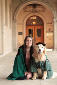 Payton Rule, wearing a green graduation gown, kneels next to her golden retriever, who wears a smaller green gown and black mortarboard graduation cap.