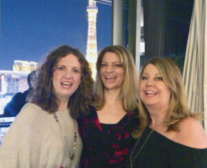 Three women smile at the camera with the neon lights of Las Vegas behind them.