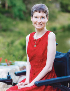 Ann Motl sits in a black wheelchair, wearing a red dress, smiling at the camera.