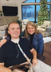 Brock Dahlke sits in a power wheelchair, smiling, with a trach vent tube attached to his neck. His mom has her arm slung around his shoulder. They are in a large, comfortable living room with a Christmas tree in the background.