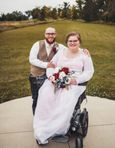 Wedding photo of Ashleigh and James Ocasio. She sits in a power wheelchair wearing a wedding dress of sheer white material layered over pink and holding a flower bouquet. He stands to her right with one arm around her shoulders, wearing a white collared shirt, pink tie, and brown vest.