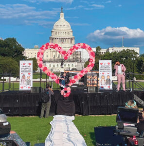 Two speakers are on an outdoor stage decorated with a heart-shaped flower arrangement and banners that say, “End the disabled marriage penalties,” with the United States Capitol building in the background.
