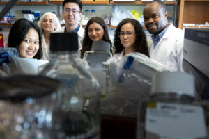 6 researchers in labcoats smile from behind a close up of lab equipment