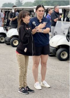 A young woman sings into a microphone held by another woman in a lot with golf carts