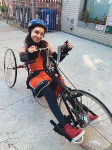 Leah Zelaya, clothed in red athletic tank top and shorts with black arm and leg sleeves and wearing a helmet, sits in a handcycle, similar to a bicycle but with two back wheels, a low reclined seat, and a chain attached to handles that rotate like pedals.