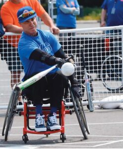 From a sitting position in a wheelchair, Jaime Zelaya swings a bat with his right arm at an incoming softball. His left hand holds the rim of the wheel as if he is prepared to move after his hit.
