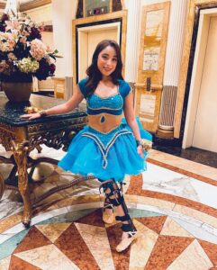 Leah, a teen with long dark brown hair and a light skin tone, stands with one hand resting on a table in a dance costume of blue short-sleeved top and tutu. On her legs and feet she wears the bilateral leg braces she uses for adaptive dance and ballet slippers.