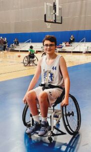 Brayden Tiernan, a teen with short brown hair, black rimmed glasses, and a light skin tone, wears white sleeveless basketball jersey and shorts and sits near the endline of a basketball court in a wheelchair with wheels that angle inward at the top.