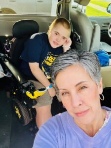 Selfie of Carol Tharp, a woman with short salt-and-pepper hair, light skin, and silver hoop earrings, with her son sitting in a power wheelchair behind her.