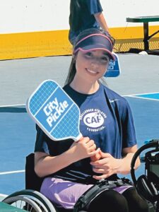 Closeup of Leah outdoors wearing a black sun visor and holding a blue paddle with white text that says City Pickle.