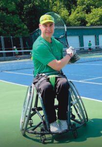 Jaime, a man wearing a yellow baseball cap and green short-sleeve shirt, poses for the camera holding a tennis racket over his left shoulder in a two-handed grip, as if about to hit a ball. He is sitting in a wheelchair with wheels that angle inward at the top on the sideline of a tennis court.