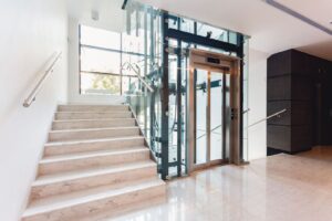 A marble stairwell with natural lighting from a large window has multiple flights of stairs that wrap around a glass elevator shaft.