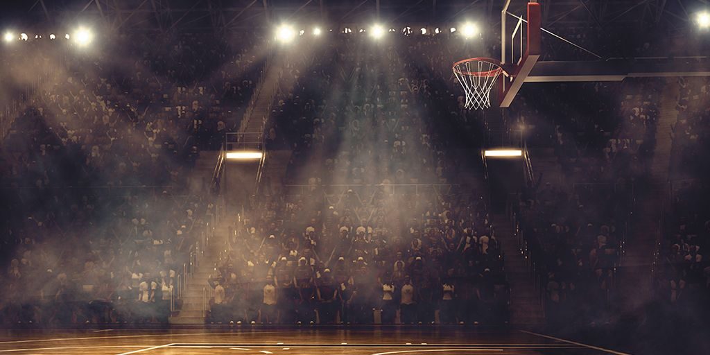 Spotlights illuminate a basketball court with no players on it and cast shadows on stands filled with fans.
