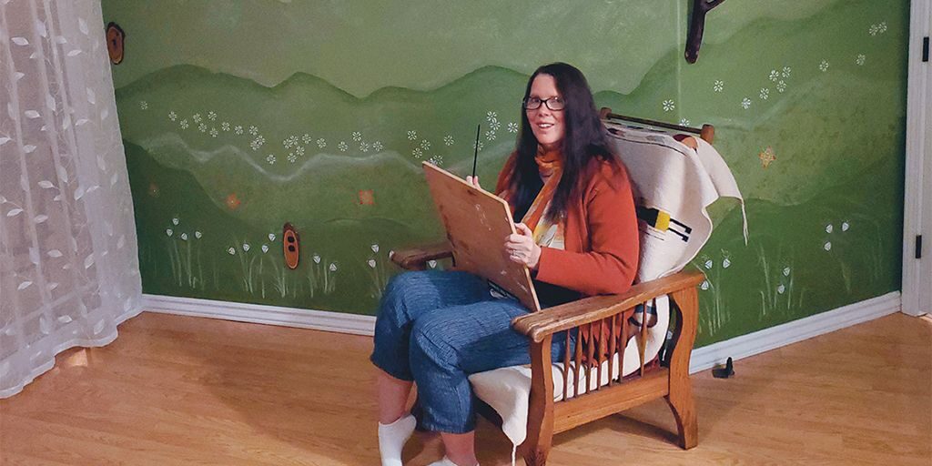 Heather Nightingale, a woman with long dark brown hair and light skin, sits in a comfortable chair next to a wall painted with a mural of rolling green hills dotted with white flowers.