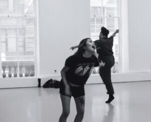 Black and white photo of Carol Alvarez in motion in a dance studio, dressed casually in a black T-shirt and shorts, swinging her arms with her head tipped back.