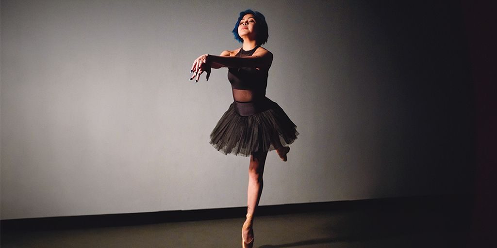 A spotlight illuminates ballerina Carol Alvarez holding an arabesque pose on pointe, wearing a black leotard and tutu.