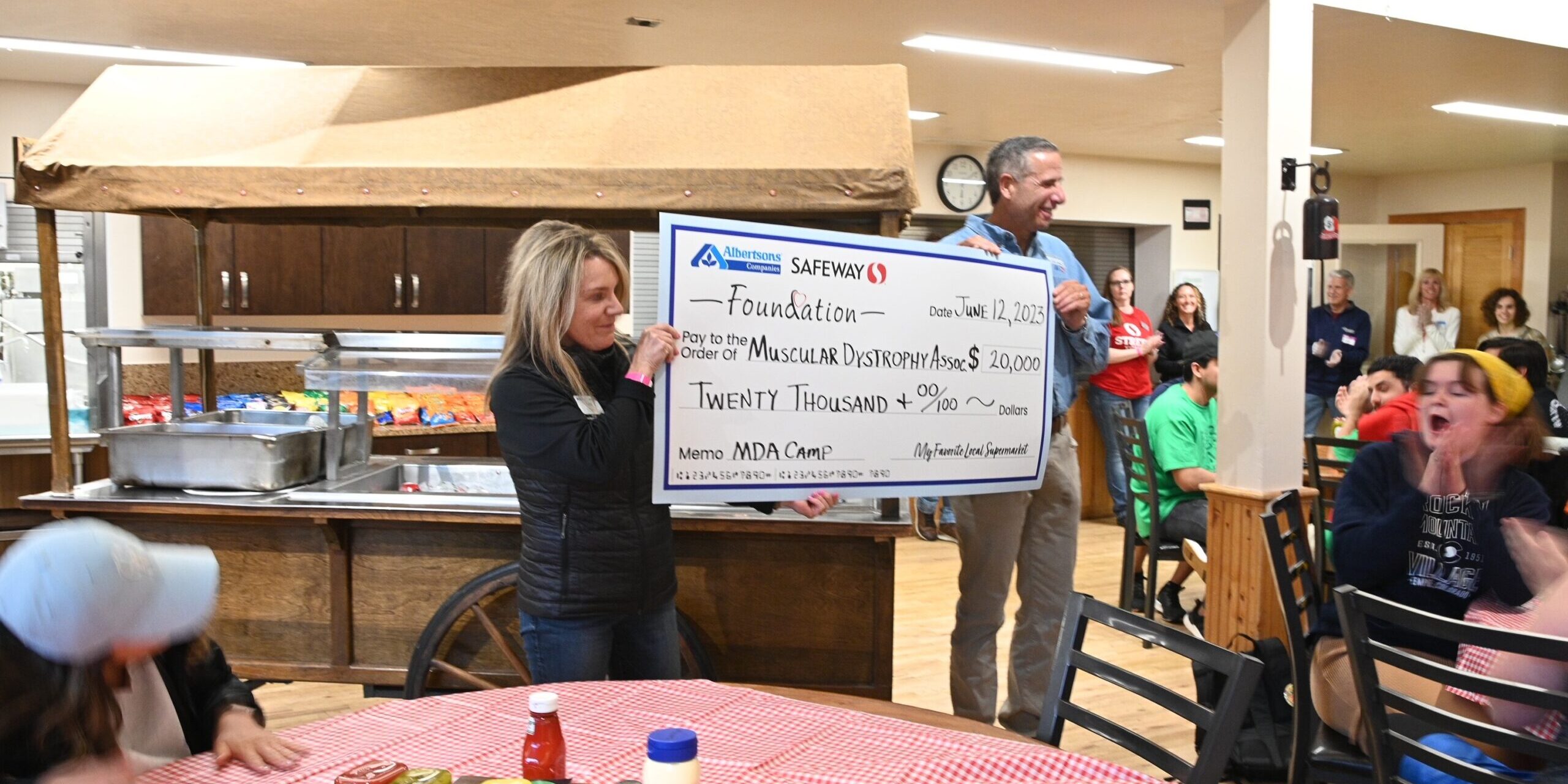 A man and woman hold a large check to donate to MDA Summer Camp in the camp cafeteria