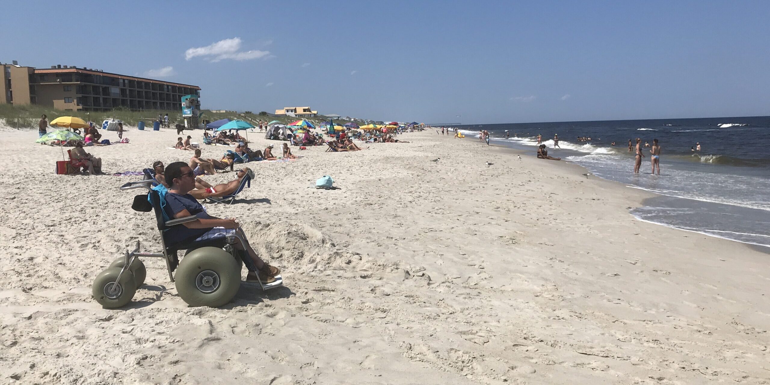 Cory Lee, a man with short, dark hair, light skin and black-rimmed glasses, enjoys an accessible beach destination in a beach wheelchair with fat tires.
