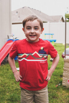 A small boy in a red shirt smiles with his hands on his hips