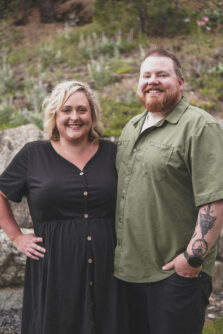 A woman in a black dress with blonde hairs smiles next to her husband, a man with a beard wearing a green shirt 