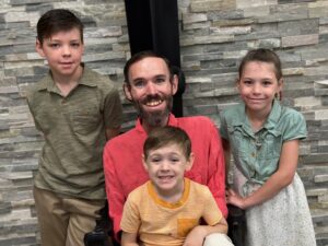 Isaac Banks, a man with light skin, short brown hair, and a beard, sits in a power wheelchair with his youngest son in his lap. His older son and daughter stand on either side of him.