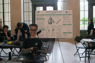 A young man wearing glasses sits in a power wheelchair in front of a presentation board about his feeding device design