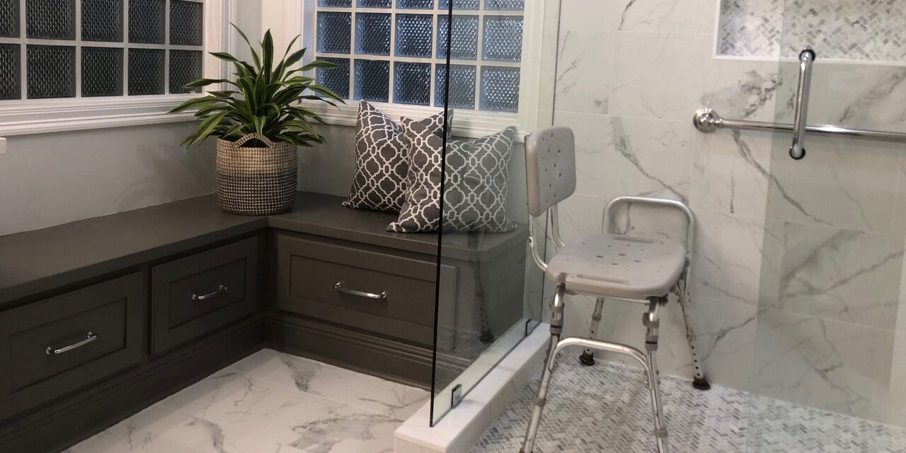 Accessible bathroom with a white marble-tiled shower with doorless entry and a gray shower chair and a brown wooden built-in bench beside it.