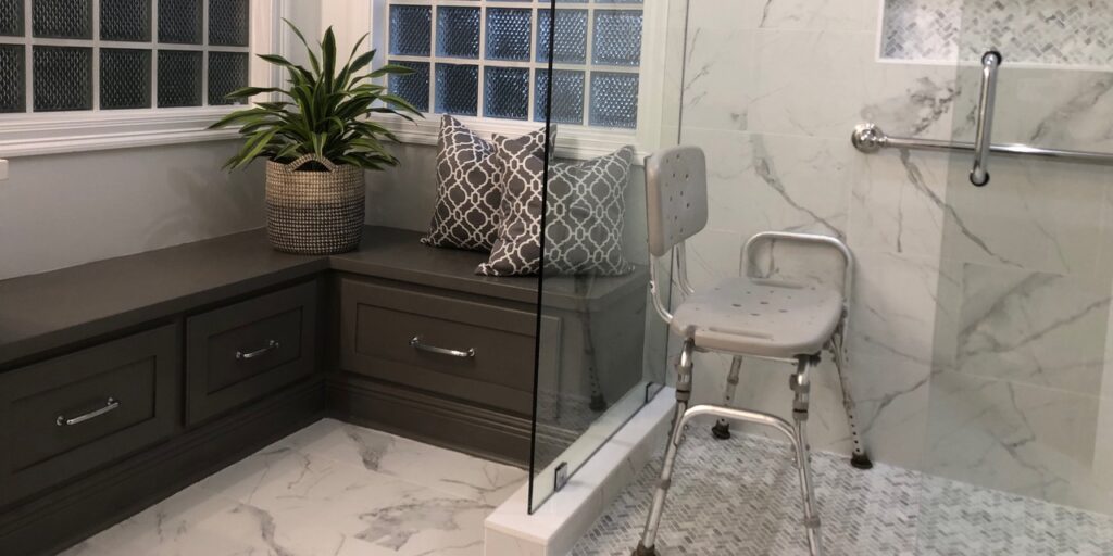 Accessible bathroom with a white marble-tiled shower with doorless entry and a gray shower chair and a brown wooden built-in bench beside it.