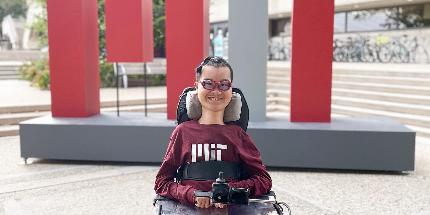 A young man in an MIT shirt and power wheelchair smiles in front of an MIT sign