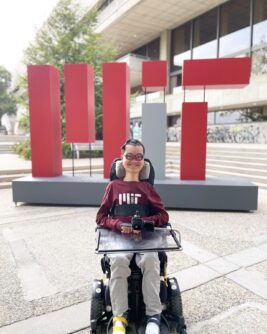 A young man sits in a power chair in front of an MIT sign