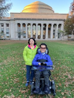 A young man in a blue winter coat and power wheelchair smiles next to a woman in green winter coat in front of a building