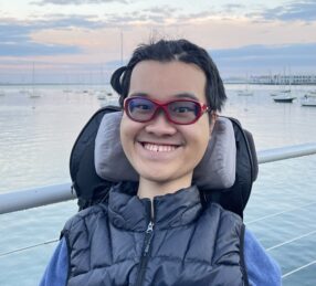 Headshot of Ben Lou, a young man wearing glasses and a winter coat in power wheelchair in front of water 