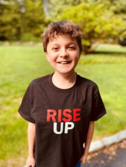 A young boy smiles outside wearing a black t-shirt that says RISE UP