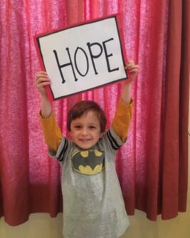 A young boy wearing a batman shirt holds up a sign that says HOPE