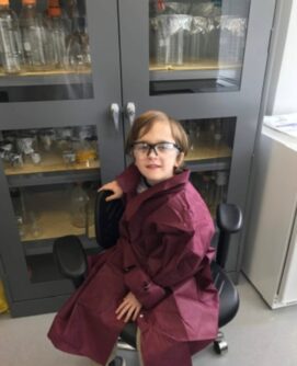 A boy wearing glasses and a lab gown smiles in front a bookshelf of beakers