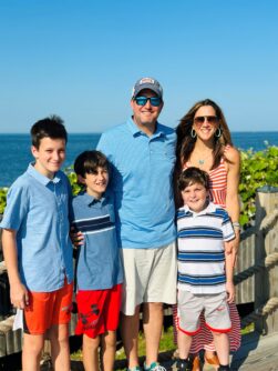 A family with a mom, dad, and three sons pose outside in front of the ocean