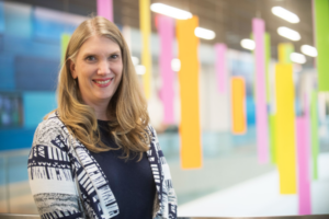 Closeup of Kathryn Mosher, a woman with light skin and long blond hair, in front of a colorful blurred background of orange, yellow, pink, and green art pieces.