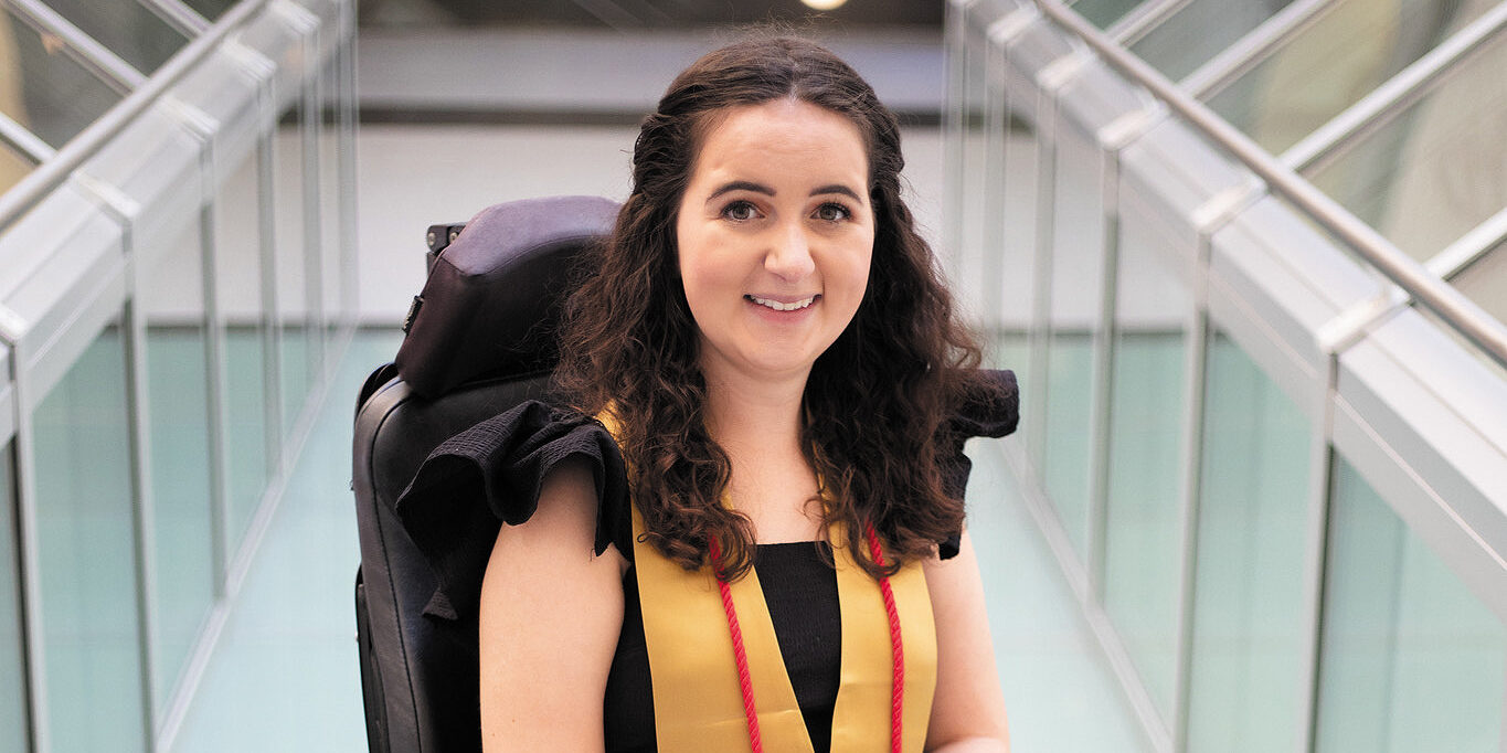 Closeup of Olivia Holler sitting in a black power wheelchair wearing a gold graduation stole with a red braided cord.