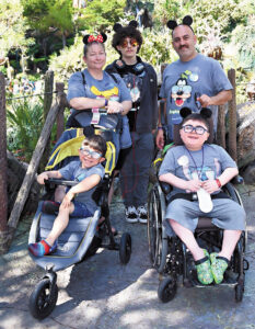 The Bedirlian family (mother, father, oldest son standing, middle son sitting in a wheelchair, and youngest son sitting in a stroller) on a Disneland trip, all wearing mouse ear headbands.